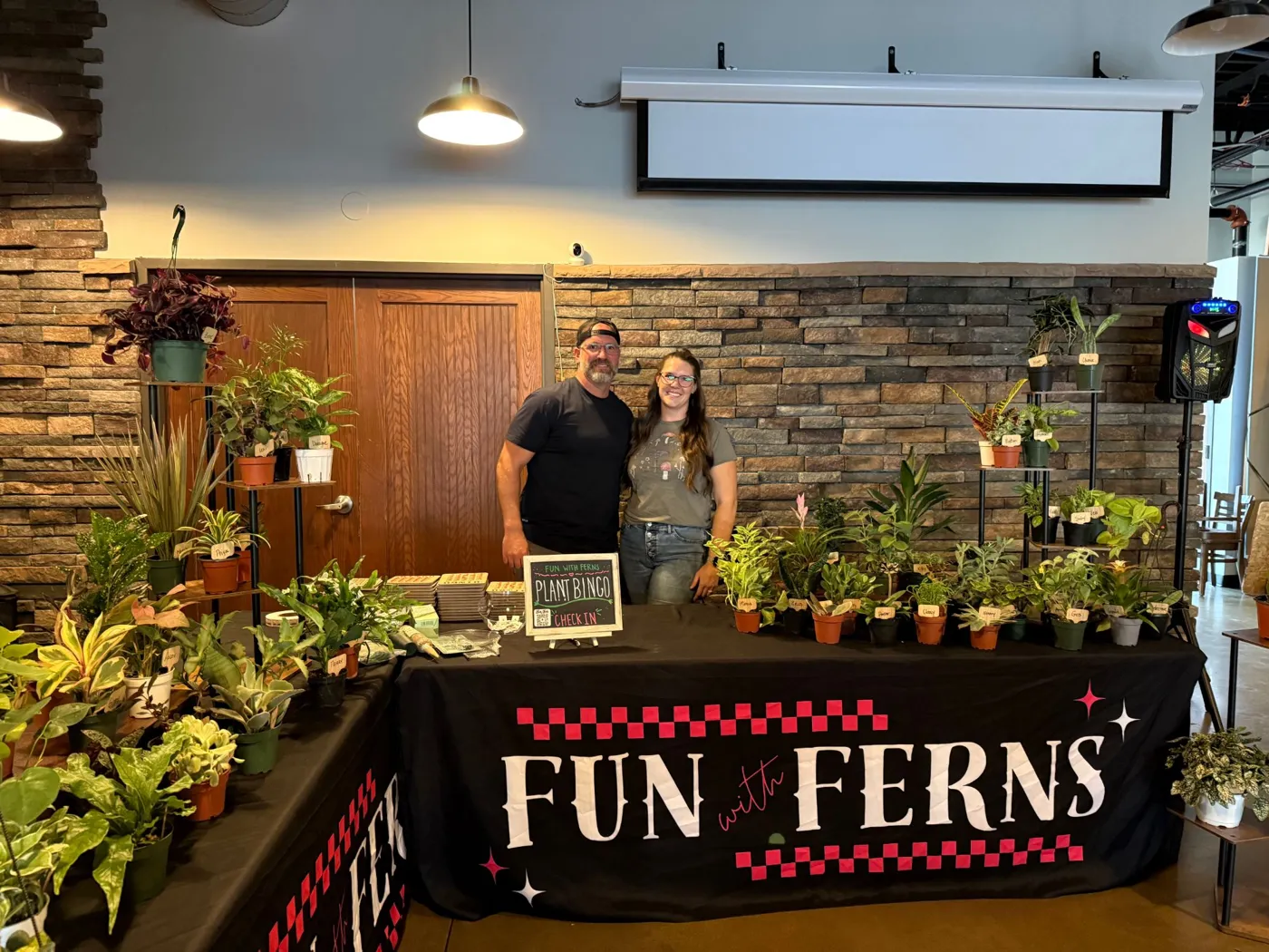 Alyssa and Michael standing behind their Fun with Ferns plant bingo table full of houseplants