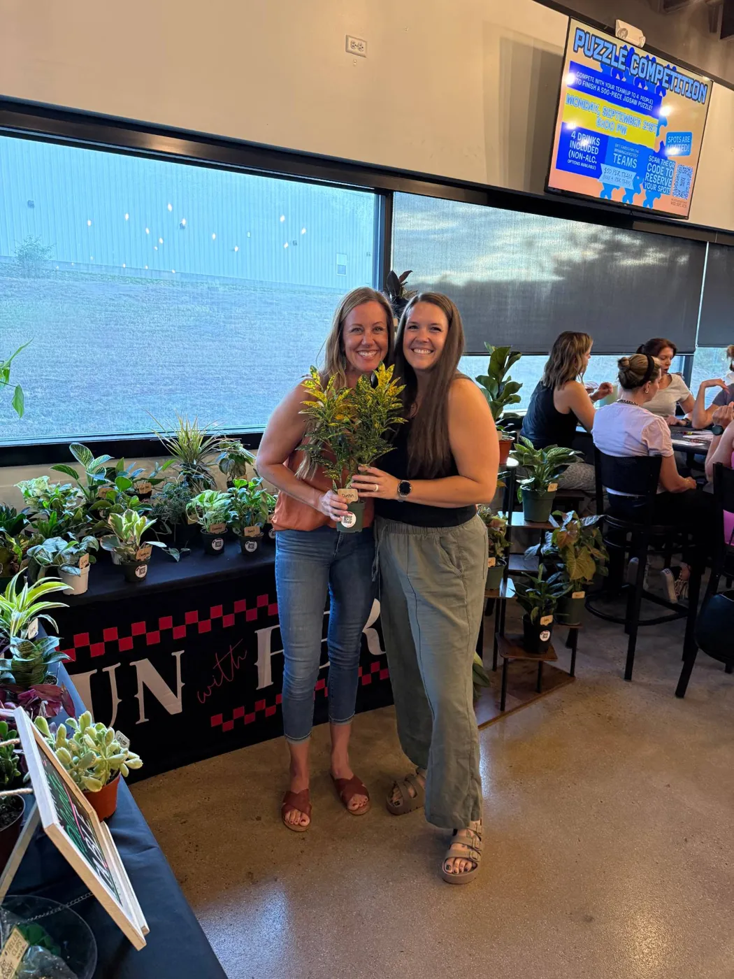 Two women smiling and holding a speckled croton in front of a table full of houseplants